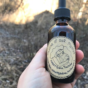 K&ouml;nnte beinhalten: Eine braune Glasflasche mit Tropfer und einem Etikett mit der Aufschrift "Medizin unserer Mutter Happy Brain Tinktur Zitronenmelisse, Ashwagandha, Mutterkraut". Das Etikett zeigt eine Cartoon-Illustration einer Person, die einen Globus h&auml;lt.