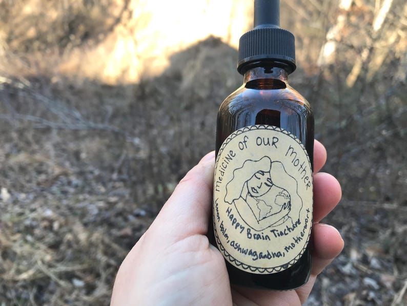 K&ouml;nnte beinhalten: Eine braune Glasflasche mit Tropfer und einem Etikett mit der Aufschrift "Medizin unserer Mutter Happy Brain Tinktur Zitronenmelisse, Ashwagandha, Mutterkraut". Das Etikett zeigt eine Cartoon-Illustration einer Person, die einen Globus h&auml;lt.