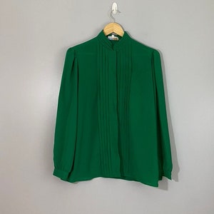 Puede incluir: Una blusa verde de manga larga con cuello alto y cierre de botones en la parte delantera. La blusa tiene pliegues en la parte delantera y está colgada de una percha de madera.