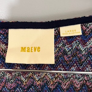 May include: A white tag with the word "MAeve" printed in gold lettering, attached to a blue and purple chevron patterned fabric. A smaller white tag with the text "LARGE MADE IN VIETNAM" is visible in the top right corner.