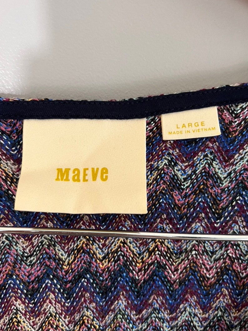May include: A white tag with the word "MAeve" printed in gold lettering, attached to a blue and purple chevron patterned fabric. A smaller white tag with the text "LARGE MADE IN VIETNAM" is visible in the top right corner.