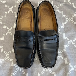 K&ouml;nnte beinhalten: Ein Paar schwarze Leder-Slipper mit schlichtem Design. Die Slipper haben eine abgerundete Spitze und einen einzigen Riemen &uuml;ber dem Spann.