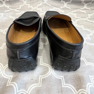 K&ouml;nnte beinhalten: Ein Paar schwarze Leder-Slipper mit braunem Leder-Innenfutter. Die Slipper haben eine Gummisohle mit einem strukturierten Muster.