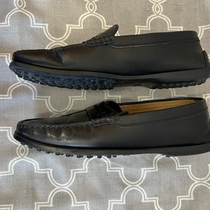 K&ouml;nnte beinhalten: Ein Paar schwarze Leder-Slipper mit Gummisohle. Die Slipper haben eine Mokassin-Zehenform und ein Quasten-Detail.