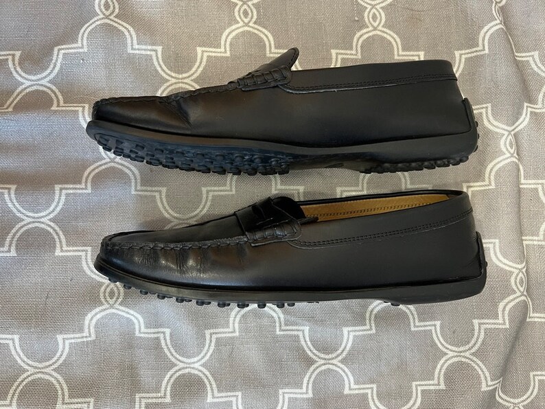 K&ouml;nnte beinhalten: Ein Paar schwarze Leder-Slipper mit Gummisohle. Die Slipper haben eine Mokassin-Zehenform und ein Quasten-Detail.