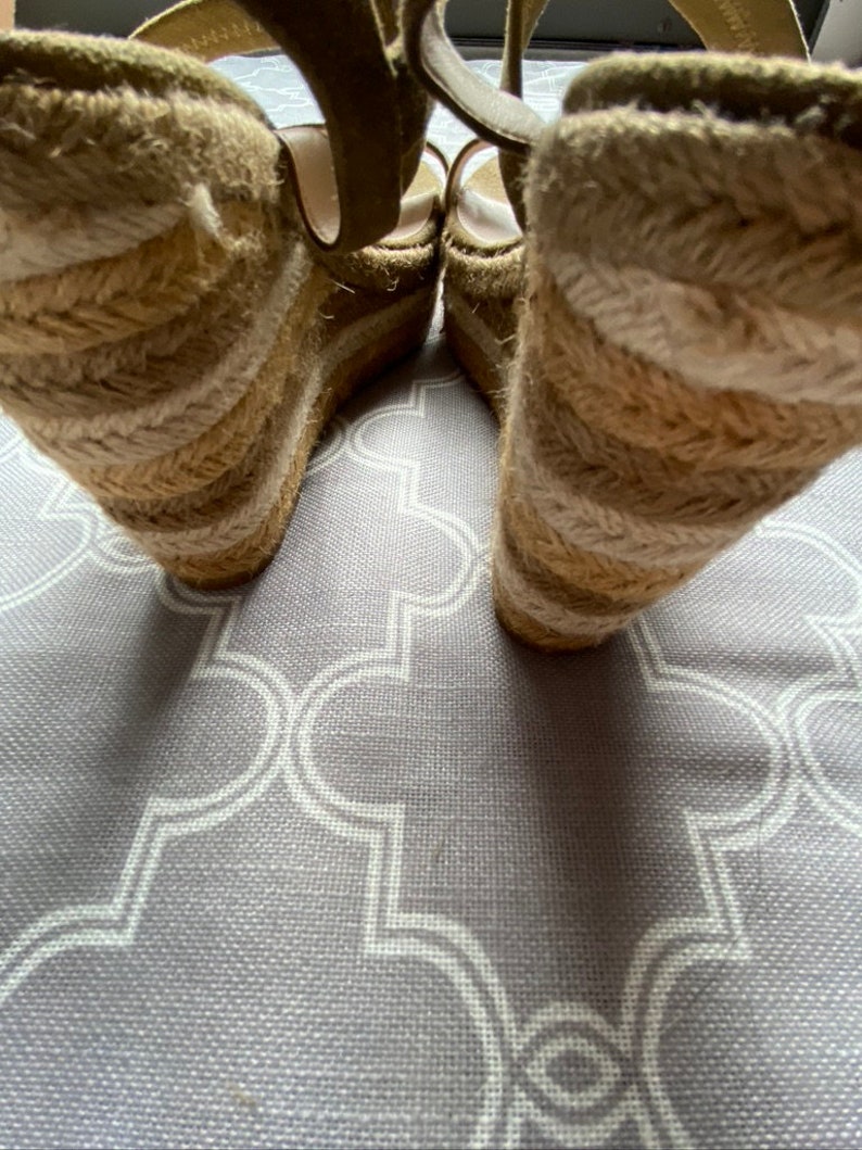May include: A pair of beige wedge sandals with a woven jute platform. The sandals have a strap that goes around the ankle and a buckle closure.