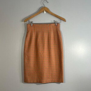 May include: A pink and green plaid skirt hanging on a wooden hanger. The skirt is knee-length and has a straight silhouette.