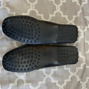 K&ouml;nnte beinhalten: Ein Paar schwarze Leder-Slipper mit einer Gummisohle. Die Sohlen haben ein strukturiertes Muster aus kleinen Kreisen.