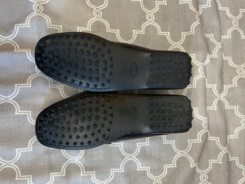 K&ouml;nnte beinhalten: Ein Paar schwarze Leder-Slipper mit einer Gummisohle. Die Sohlen haben ein strukturiertes Muster aus kleinen Kreisen.