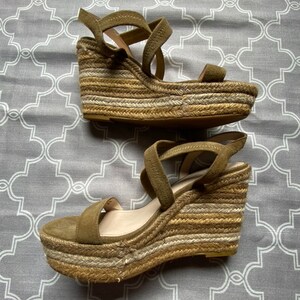May include: A pair of olive green suede wedge sandals with a woven jute platform. The sandals have a criss-cross strap design and an ankle strap.