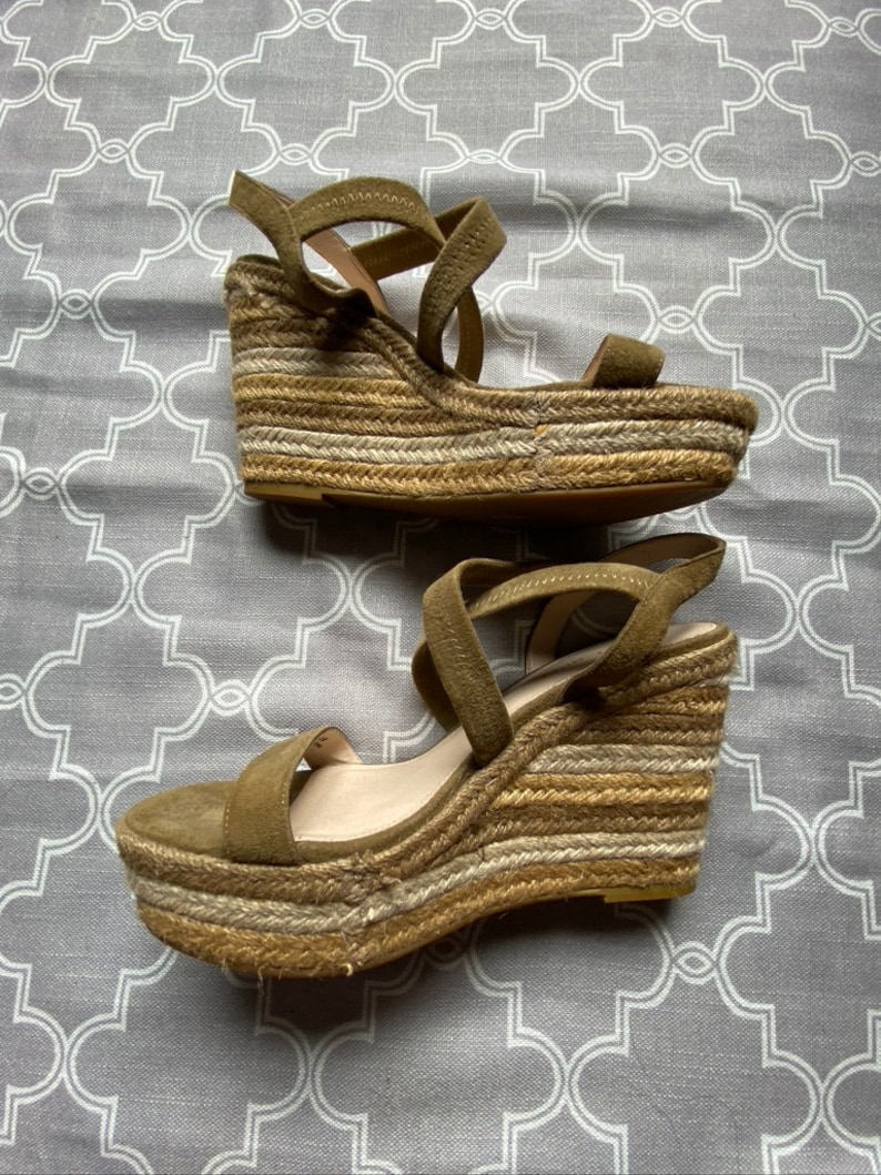 May include: A pair of olive green suede wedge sandals with a woven jute platform. The sandals have a criss-cross strap design and an ankle strap.