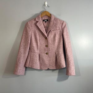 May include: A dusty rose blazer featuring a textured fabric, a notched lapel, and three brown buttons down the front. The garment includes two front waist pockets and a small chest pocket, with a tailored fit and long sleeves hanging on a hanger.