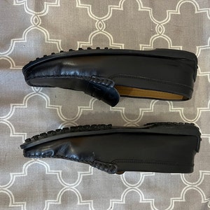 K&ouml;nnte beinhalten: Ein Paar schwarze Leder-Slipper mit einem gestickten Design an der Spitze. Die Slipper haben eine Gummisohle und liegen auf einer gemusterten Oberfl&auml;che.