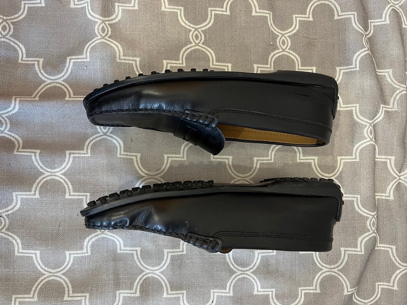 K&ouml;nnte beinhalten: Ein Paar schwarze Leder-Slipper mit einem gestickten Design an der Spitze. Die Slipper haben eine Gummisohle und liegen auf einer gemusterten Oberfl&auml;che.