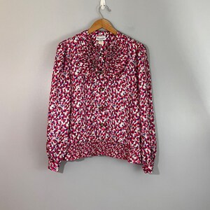 Può includere: Una blusa a maniche lunghe con motivo bianco, rosa e blu, con colletto a volant e chiusura con bottoni. La blusa ha una cintura elastica arricciata.