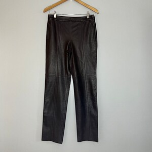 May include: Dark brown leather trousers hanging on a wooden hanger. The trousers have a straight leg cut and visible seams down the front and back. The leather appears smooth and has a slight sheen. The trousers are displayed against a plain, light-coloured background.