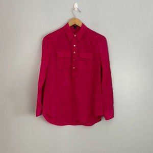 May include: A long-sleeved, pink button-down shirt with two chest pockets. The shirt has a collared neckline and gold buttons.