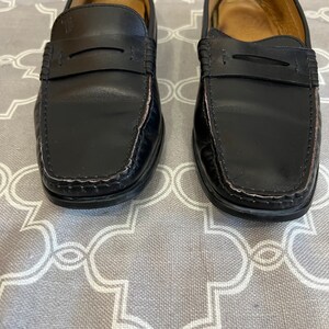 K&ouml;nnte beinhalten: Ein Paar schwarze Leder-Slipper mit schlichtem Design. Die Slipper haben eine abgerundete Spitze und eine Naht am Rand.