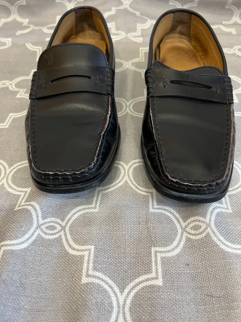 K&ouml;nnte beinhalten: Ein Paar schwarze Leder-Slipper mit schlichtem Design. Die Slipper haben eine abgerundete Spitze und eine Naht am Rand.