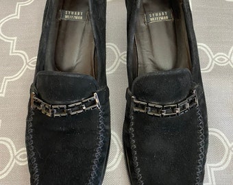May include: Black suede loafers with a chain detail across the front. The loafers have a stitched edge and the brand name "Stuart Weitzman" is printed on the insole.