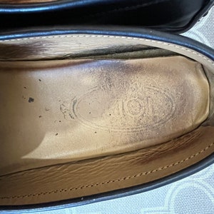 K&ouml;nnte beinhalten: Nahaufnahme des Innenraums eines schwarzen Lederschuhs mit Schnallen. Die Innensohle ist aus braunem Leder mit einem runden Logo, auf dem "TOD'S" in einer stilisierten Schrift steht.
