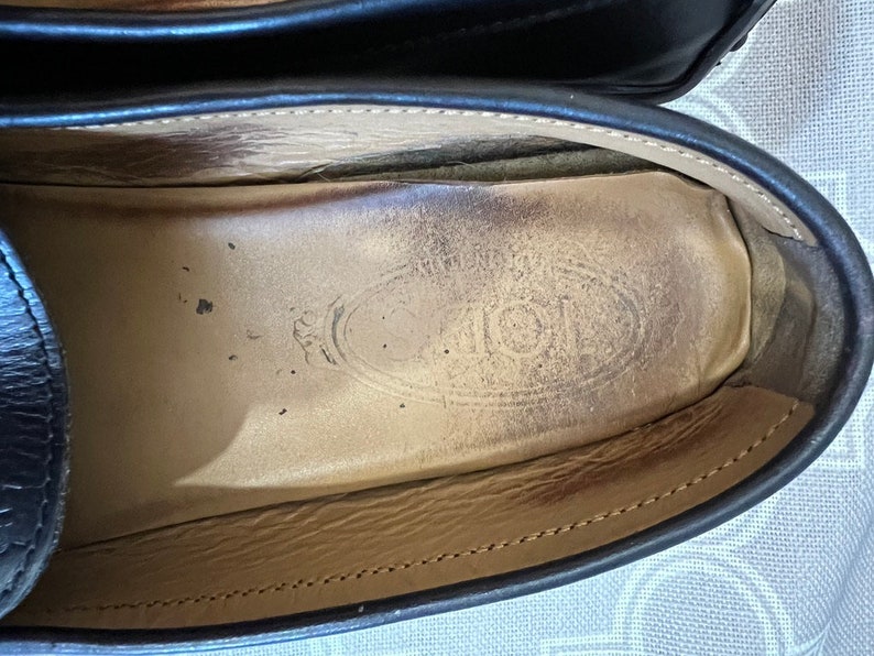 K&ouml;nnte beinhalten: Nahaufnahme des Innenraums eines schwarzen Lederschuhs mit Schnallen. Die Innensohle ist aus braunem Leder mit einem runden Logo, auf dem "TOD'S" in einer stilisierten Schrift steht.