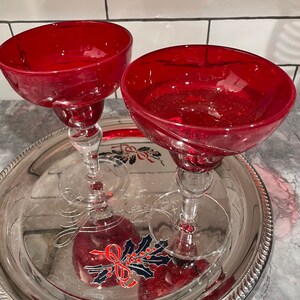 May include: Two red glass margarita glasses with a sparkly finish sit on a silver tray with a holiday design.