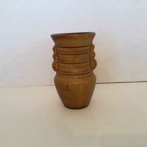 May include: A small, brown ceramic vase with two handles. The vase has a textured surface and is decorated with horizontal lines.