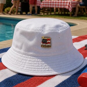 May include: White bucket hat with a patriotic pin featuring red, white, and blue beads arranged in a flag design. The hat is on a red, white, and blue striped towel near a pool, suggesting a summer theme.