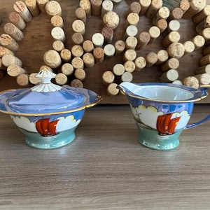 Puede incluir: Un juego de azucarero vintage con tapa y jarra de porcelana. El juego presenta un diseño azul claro y dorado con un motivo de velero rojo. La jarra tiene un asa y el azucarero tiene una tapa decorativa.