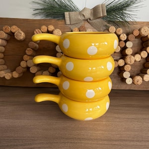 May include: A stack of four yellow ceramic soup bowls with handles, each adorned with white polka dots. The bowls are stacked on top of each other, creating a visually appealing arrangement. The background includes a wooden surface and decorative elements.