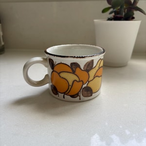 Pode incluir: Uma pequena caneca de cerâmica com base creme e borda marrom. A caneca é decorada com desenhos florais laranja e amarelo e tem uma pequena alça. Um vaso de plantas branco está no fundo.