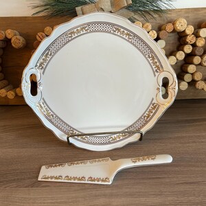 May include: A white ceramic serving platter with gold trim and handles. The platter features a decorative floral pattern and is displayed on a wooden surface with a cork design.