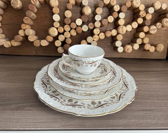 May include: A set of four white porcelain plates with a gold rim and a delicate floral pattern. The plates are stacked on top of each other, with a small white teacup with a gold rim and floral pattern on top.