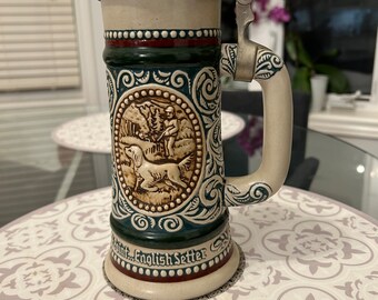 May include: A ceramic beer stein with a pewter lid and a handle. The stein features a hunting scene with a dog and a hunter. The text "Point... English Setter" is printed on the bottom of the stein.