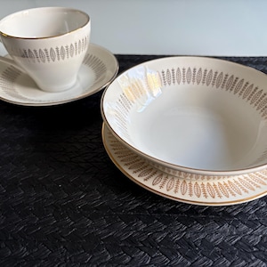 Peut inclure: Un service à thé blanc avec un motif de feuilles dorées, accompagné d'un bol et d'une assiette assortis. L'ensemble est posé sur une surface noire texturée, mettant en valeur un design classique et élégant. Le bol et l'assiette mesurent environ 20 cm de diamètre.