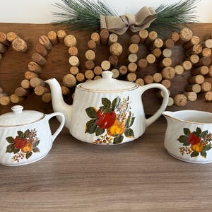 Peut inclure: Service à thé en céramique blanche comprenant une théière, un sucrier et une crémière. Chaque pièce est ornée d'un motif de fruits et de fleurs rouges, jaunes et vertes. L'ensemble est bordé d'or et posé sur une surface en bois.