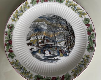 May include: A white ceramic plate with a red, green, and yellow border featuring a winter scene with people ice skating on a frozen pond. The scene includes a small wooden house with a water wheel and trees covered in snow.