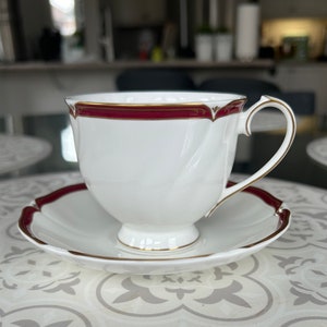 May include: A white teacup and saucer set with a burgundy and gold trim. The cup has a delicate, fluted design.