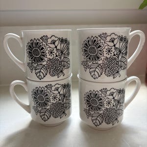 May include: Four white ceramic mugs arranged in two stacks of two. Each mug features a black line art illustration of a floral bouquet containing sunflowers, clover-like blossoms, and various leaves. The mugs have a simple handle and base.