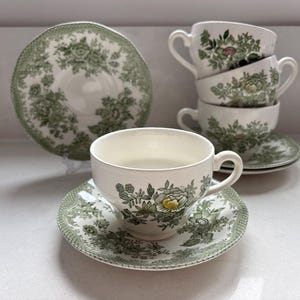 Puede incluir: Un juego de tazas y platillos vintage. Las tazas y los platillos son blancos con un estampado floral verde. El juego incluye una pila de cuatro tazas, un platillo y un plato grande. Las tazas tienen asa y están colocadas sobre platillos a juego.