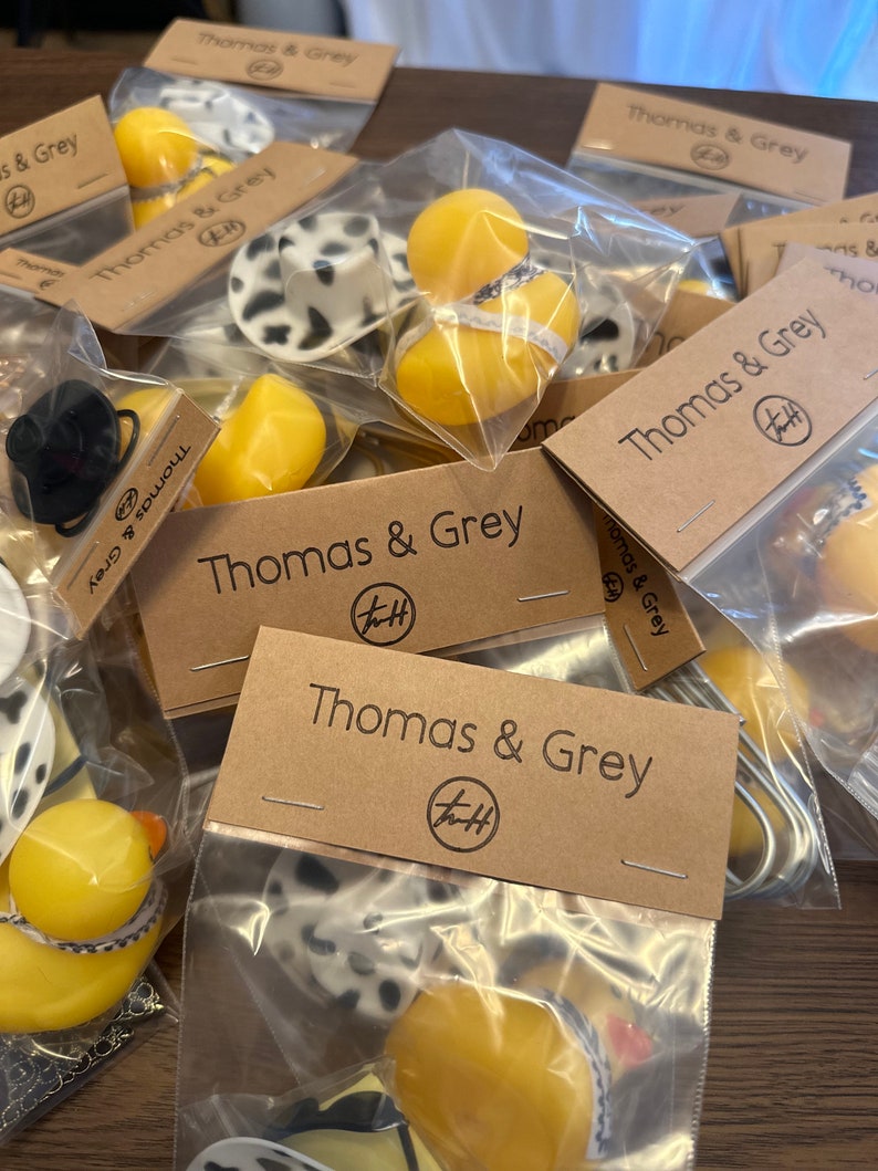 May include: A collection of small, yellow rubber duckies packaged in clear plastic bags with brown paper tags. The tags have the text "Thomas & Grey" and a logo with the letters "T&H".