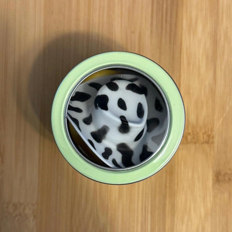 May include: A black and white cow print hat inside a green tin can with a clear lid.