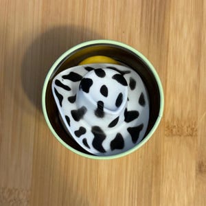 May include: A black and white cow print hat in a green and brown container.