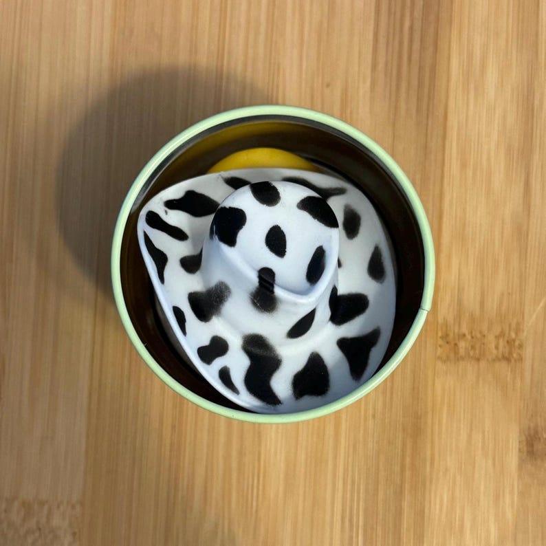 May include: A black and white cow print hat in a green and brown container.