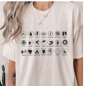 May include: White t-shirt with black circular icons and text. The icons represent different powers, including growth, shade, teleporter, and more. The text below each icon names the power.