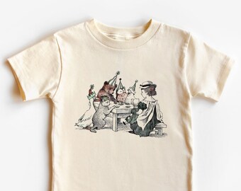 Op de afbeelding: Een crèmekleurig T-shirt met een vintage illustratie van een meisje en dieren die een theepartij houden. Het meisje draagt een hoed en een jurk. De dieren zijn een beer, een kat en een gans. Alle dieren dragen feestmutsen.