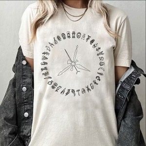 May include: A white t-shirt featuring a black and white graphic design of a circle with symbols and a starburst in the centre.