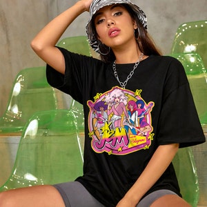 Może przedstawiać: Czarny T-shirt z grafiką przedstawiającą zesp&oacute;ł kresk&oacute;wkowy Jem and the Holograms. Członkowie zespołu są ubrani w kolorowe stroje i trzymają gitary. Koszulka ma na sobie biało-czarną czapkę w paski.