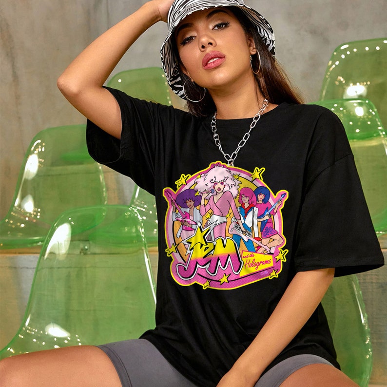 Może przedstawiać: Czarny T-shirt z grafiką przedstawiającą zesp&oacute;ł kresk&oacute;wkowy Jem and the Holograms. Członkowie zespołu są ubrani w kolorowe stroje i trzymają gitary. Koszulka ma na sobie biało-czarną czapkę w paski.